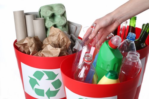 A person's hand is placing a clear plastic bottle into a red recycling bin, which is positioned among another bin filled with various empty bottles, jars, and containers. The bins are decorated with green recycling symbols on a white background, indicating their purpose for waste separation. The waste inside the bins includes crumpled brown paper, cardboard tubes, and different plastic bottles, some with caps attached, and others without. The items are organized in the bins, with the bottles in the foreground and paper materials stacked behind. The scene appears to be set on a bright day, with a clean, urban or residential outdoor setting suggested by the plain white background. This visual relates to waste collection and recycling services, specifically those offered by Garden Clearance Hampstead Heath, which handle rubbish removal and proper waste disposal in the local area. The focus on different recyclable materials underscores a professional approach to waste management and environmental responsibility typical of such services within Hampstead Heath and surrounding NW postcode areas.