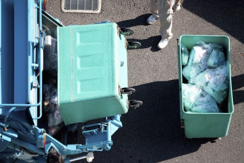 An overhead view of a waste collection scene showing a large, light green, wheeled refuse bin open at the top, filled with plastic waste bags and discarded packaging materials, positioned on a dark asphalt surface. Next to it, a matching green bin is also filled with similar waste items, with clear plastic bags containing rubbish visible inside. A person partially visible, dressed in light-coloured clothing and white shoes, stands near the bins, hinting at a rubbish removal or garden clearance activity. The background includes a section of a blue waste vehicle with mechanical arms, indicating an urban or suburban environment, possibly within Hampstead Heath or adjoining areas in north London. The image relates to local rubbish collection services and garden clearance for properties in the target postcode area.