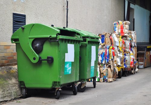 In the foreground, there are two large, green wheeled rubbish bins with black lids, positioned against a beige external wall of a building, with one bin slightly overlapping the other. The bins are made of durable plastic, featuring textured surfaces and visible locking mechanisms on the lids. Behind the bins, there are several tightly packed bundles of flattened cardboard boxes, variously coloured with some displaying blue, red, and brown tones, stacked neatly on the ground along the side of a paved driveway. The cardboard bundles show signs of wear, with creases and torn edges, indicating they are prepared for disposal or recycling. In the background, part of a modern building with large glass windows and a black vertical element is visible on the right, suggesting an urban or suburban environment typical of Hampstead Heath. The lighting is natural and diffuse, casting minimal shadows, highlighting the textures of the cardboard and the plastic surfaces of the bins, in a setting consistent with waste collection or rubbish removal services carried out by Garden Clearance Hampstead Heath.