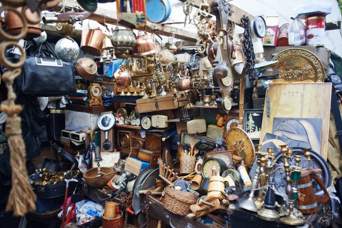 A cluttered outdoor display of various household and decorative items, including metal and wooden objects such as lamps, vases, picture frames, and miscellaneous bric-a-brac, arranged on tables and hanging from a structure. The assortment features a mix of copper, brass, and painted finishes, with some items showing signs of age or wear, indicating a second-hand or vintage collection. The background suggests an outdoor market or yard setting, with overlapping objects creating a dense, eclectic visual. The scene captures the complexity and variety of items typically encountered in rubbish clearance or household decluttering projects in areas like Hampstead Heath, where Garden Clearance Hampstead Heath offers such services. The lighting is diffuse, providing even visibility across the assortment of objects, emphasizing their textures and finishes. This detailed image of miscellaneous household items reflects the type of clutter handled by rubbish removal specialists during garden or property clearance in the local area, aiding individuals or businesses in clearing out unwanted possessions efficiently and professionally.