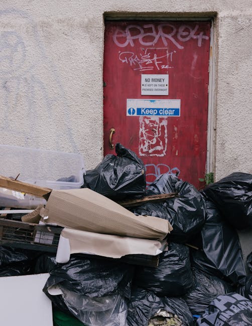 A white waste collection truck with an open rear compactor is parked on a narrow urban street lined with multi-story residential buildings featuring beige and grey facades, some showing signs of weath
